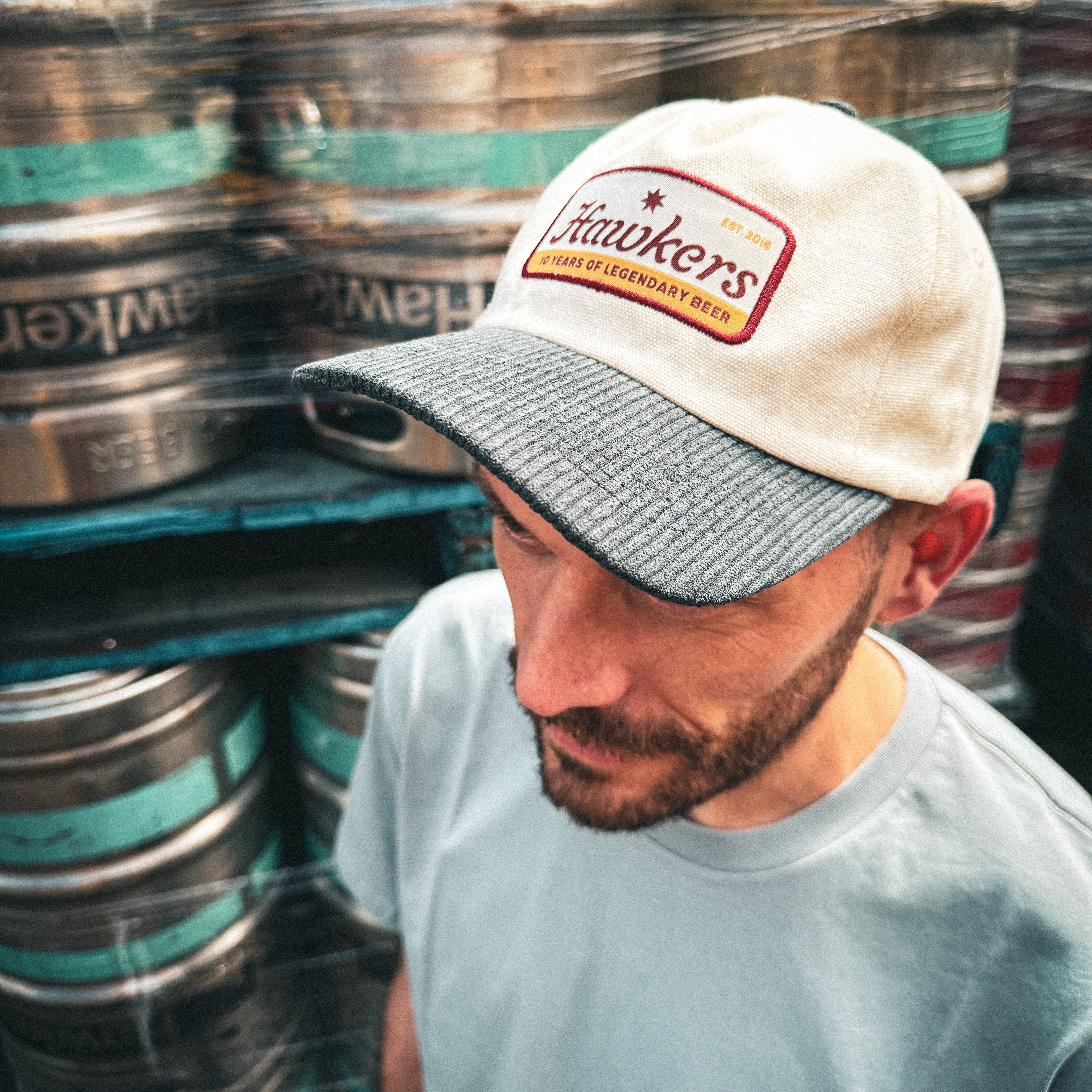 Man wearing a cap and t-shirt with 'Hawkers' branding in front of stacked kegs.